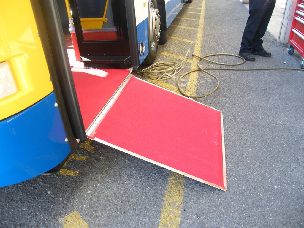 A red fold-out boarding ramp lowered from a bus doorway onto the kerb