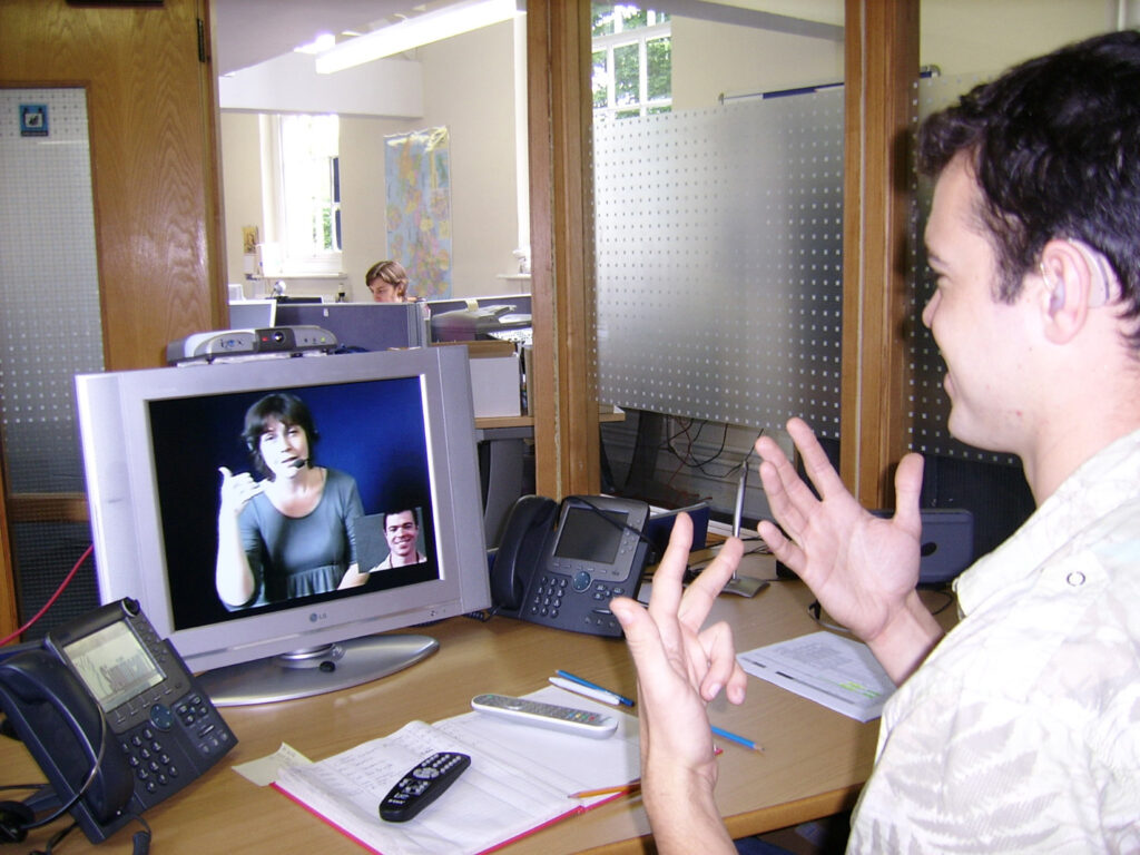 A deaf man signing to an interpreter on a video relay screen at his desk