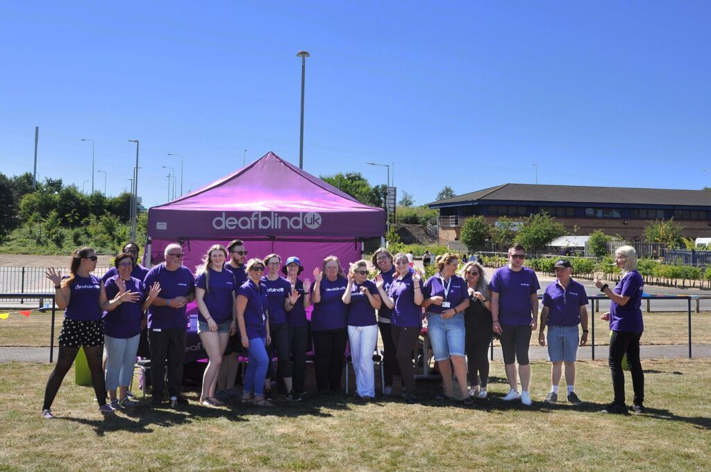 Members of Deafblind UK gathered around a purple branded gazebo at an outdoor celebration