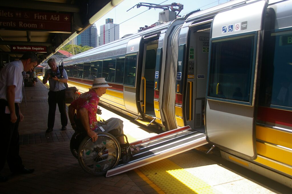 A wheelchair user rolling down a portable ramp from a train onto the platform while staff stand by