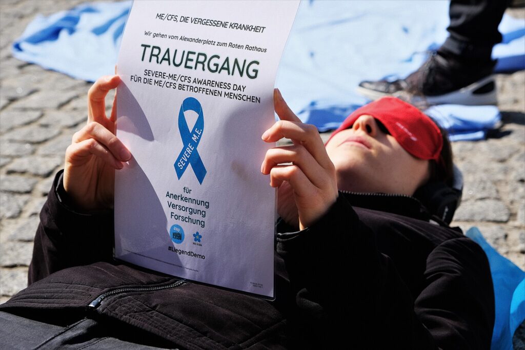 A protester lying on the ground wearing an eye mask, holding a sign with a blue ribbon for Severe ME/CFS Awareness Day