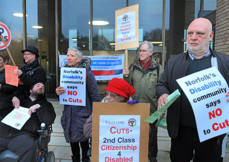 Disabled campaigners, one in a wheelchair, hold placards outside Norfolk County Hall reading Norfolk's disability community says no to cuts